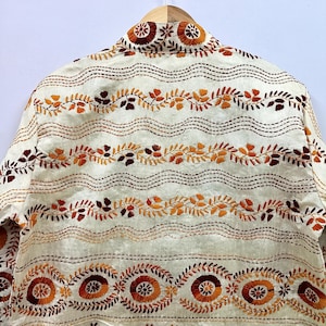 May include: An off-white jacket featuring horizontal rows of intricate embroidery in shades of orange, rust, and dark brown. The design includes repeating wavy lines, leafy vine patterns, and circular floral motifs across the fabric surface.