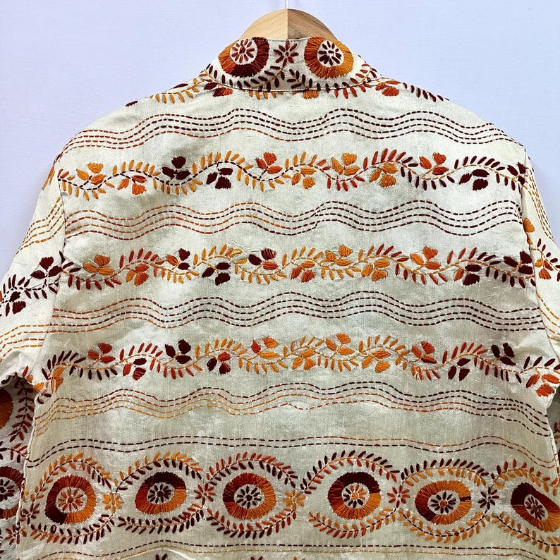 May include: An off-white jacket featuring horizontal rows of intricate embroidery in shades of orange, rust, and dark brown. The design includes repeating wavy lines, leafy vine patterns, and circular floral motifs across the fabric surface.
