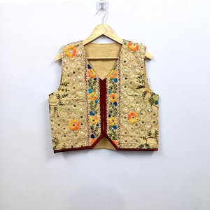 May include: A sleeveless beige waistcoat featuring intricate embroidery with orange, yellow, and blue floral patterns. The garment is adorned with small circular mirrors throughout the fabric and is finished with a dark red trim along the front edges.