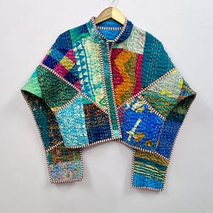Op de afbeelding: Een patchwork jasje met een opstaande kraag en een knoopsluiting. De jas is gemaakt van verschillende stoffen lappen in tinten blauw, groen, oranje en roze, met witte en rode stiksels. De jas hangt aan een houten kleerhanger.