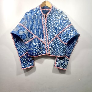 May include: A blue and white patchwork jacket with a stand-up collar and short sleeves. The jacket features various printed patterns and is trimmed with a pink border. It is hanging on a wooden hanger.