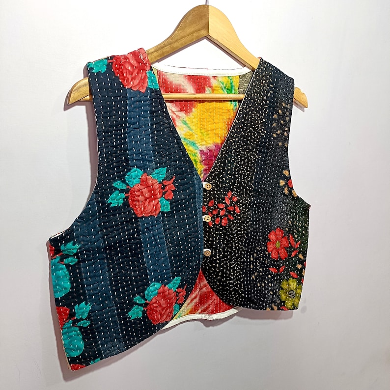 Op de afbeelding: Een omkeerbaar vest met een bloemenpatroon. De ene kant heeft een donkerblauwe achtergrond met verticale strepen en rode rozenontwerpen. De andere kant heeft een zwarte achtergrond met een bloemenpatroon. Het vest is geknoopt en hangt aan een houten hanger.