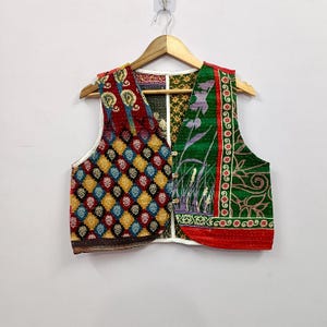 May include: A sleeveless vest featuring a patchwork design with a variety of colorful patterns. The left side displays a diamond motif in red, yellow, and blue, while the right side features green fabric with floral and botanical prints. Three buttons.