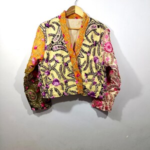 Könnte beinhalten: Eine kurze, bestickte Jacke in verschiedenen Farben. Die Jacke hat eine gelbe Basis mit lila Blumenstickerei, orangefarbenen Besatz und Ärmel mit olivgrünen und rosa Blumenmustern. Sie hängt an einem Holzbügel.
