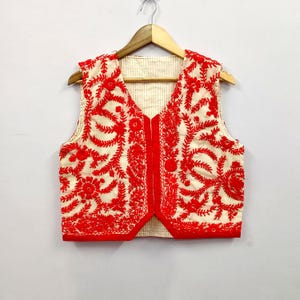 Puede incluir: Chaleco color crema con intrincado bordado floral rojo. La prenda sin mangas presenta un cuello en V y una parte delantera abotonada. El bordado cubre la parte delantera y trasera del chaleco, creando un diseño detallado y texturizado.