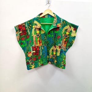 Könnte beinhalten: Eine ärmellose, gesteppte Jacke in leuchtendem Grün mit einem sich wiederholenden Muster aus Figuren und Blumenmotiven in Rot, Gelb und Blau. Das Kleidungsstück hat einen Kragen, eine offene Vorderseite und sichtbare weiße Steppnähte.