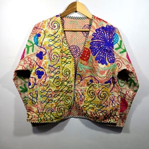 May include: An embroidered jacket with a V-neck design. The jacket has a beige base with detailed patterns in blue, yellow, pink, and green. The jacket is hanging on a wooden hanger.