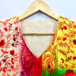 May include: A V-neck top featuring a vibrant color-blocked design with sections of cream, pink, yellow, and green. The fabric is adorned with intricate red and orange floral and paisley embroidery throughout, and the interior lining is beige striped.