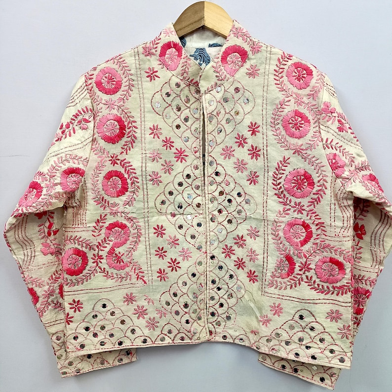 May include: An off-white jacket featuring intricate pink floral embroidery and small circular mirrors. The garment has a mandarin collar, long sleeves, and a front opening, with decorative stitching patterns covering the entire surface.