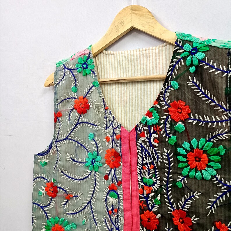 K&ouml;nnte beinhalten: Eine &auml;rmellose Weste mit V-Ausschnitt und einem zweifarbigen Design, bestehend aus einer hellgrauen und einer dunkelgrauen Seite. Das Kleidungsst&uuml;ck ist mit leuchtend orangefarbenen, gr&uuml;nen und blauen Blumenstickereien sowie einer rosa Mittelnaht verziert.