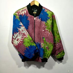 Könnte beinhalten: Blumenmuster-Bomberjacke mit schwarzem Kragen, Bündchen und Saum. Die Jacke hat ein Muster aus großen blauen, grünen und weißen Blumen auf einem malvenfarbenen Hintergrund. Sie hat einen durchgehenden Reißverschluss und zwei Taschen.