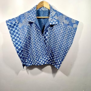 May include: A blue and white patterned wrap top with a V-neck and short sleeves. The fabric features a repeating circular and dot design. The top is hanging on a wooden hanger against a plain background.