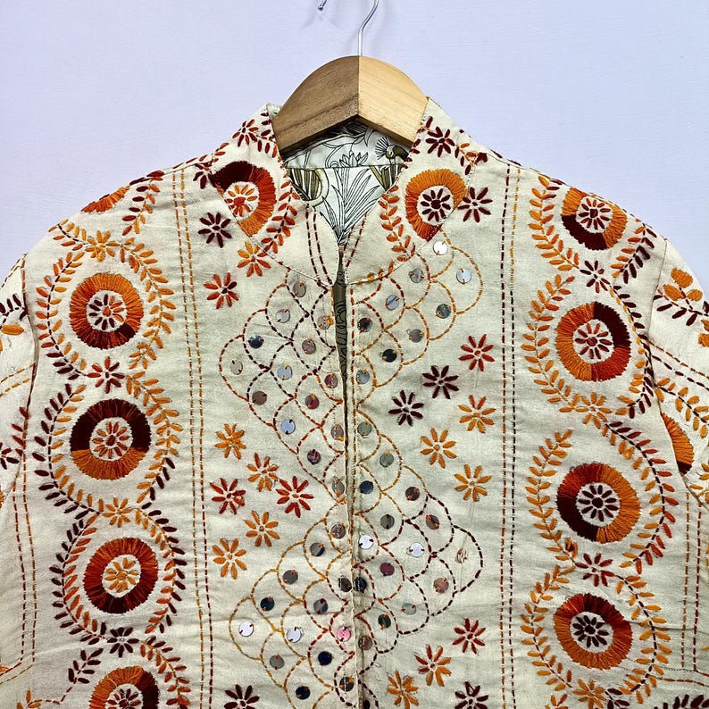 May include: An off-white jacket featuring intricate embroidery in shades of orange, rust, and brown. The garment displays circular floral patterns, vertical stitched lines, and small circular mirrors embedded within a scalloped design.