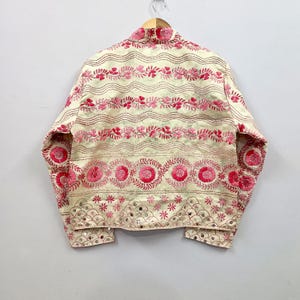 May include: A cream-colored jacket featuring horizontal bands of pink floral embroidery, wavy lines, and circular motifs. The lower section includes a scalloped pattern with small reflective mirror accents integrated into the fabric design.