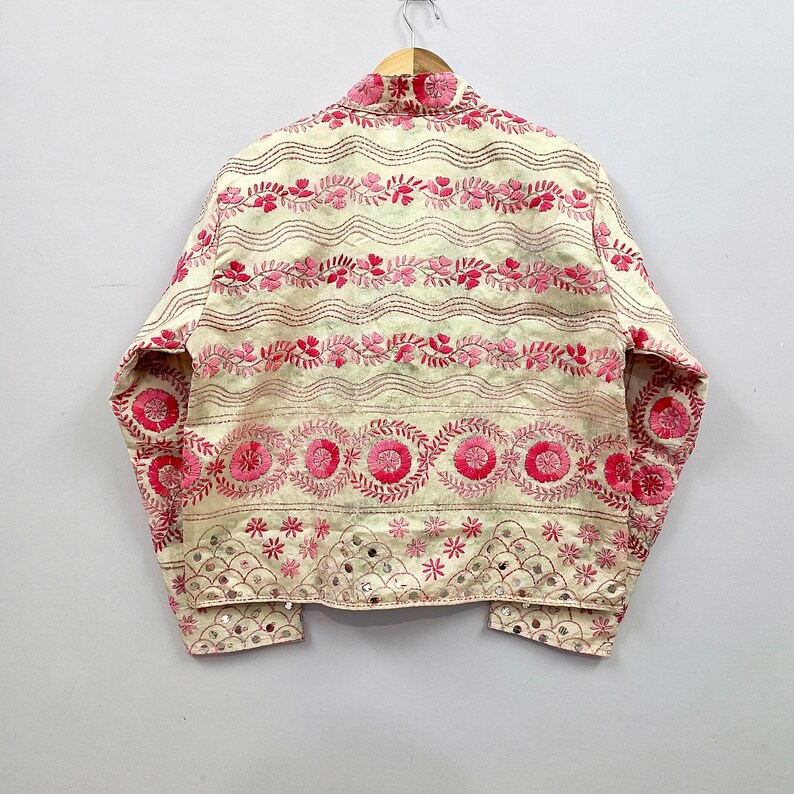 May include: A cream-colored jacket featuring horizontal bands of pink floral embroidery, wavy lines, and circular motifs. The lower section includes a scalloped pattern with small reflective mirror accents integrated into the fabric design.