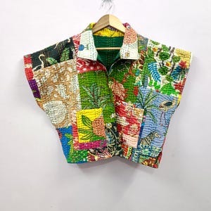 Pode incluir: Uma camisa com gola patchwork com uma vibrante mistura de cores e padrões. A camisa apresenta uma variedade de retalhos de tecido com motivos botânicos e animais, incluindo palmeiras, zebras e flamingos. A camisa está pendurada em um cabide de madeira.