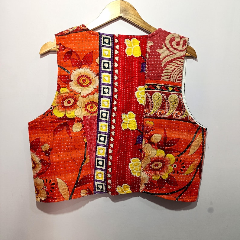 Op de afbeelding: Een patchwork vest met een levendig bloemenontwerp. Het vest heeft een mix van rode, oranje en gele stoffen met ingewikkelde stiksels. Het vest is mouwloos en hangt aan een houten hanger.