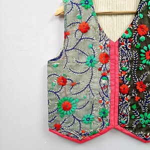 K&ouml;nnte beinhalten: Eine &auml;rmellose Weste mit V-Ausschnitt und einem lebendigen Blumenstickmuster. Die linke Seite ist hellgrau, die rechte Seite dunkelgrau, beide akzentuiert mit leuchtend orangefarbenen und t&uuml;rkisfarbenen Bl&uuml;ten, blauen Ranken und pinkem Saum.