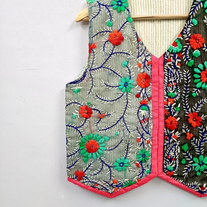 K&ouml;nnte beinhalten: Eine &auml;rmellose Weste mit V-Ausschnitt und einem lebendigen Blumenstickmuster. Die linke Seite ist hellgrau, die rechte Seite dunkelgrau, beide akzentuiert mit leuchtend orangefarbenen und t&uuml;rkisfarbenen Bl&uuml;ten, blauen Ranken und pinkem Saum.