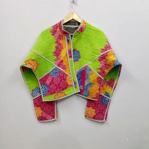 May include: A cropped jacket featuring a vibrant patchwork design with lime green, magenta, yellow, and blue floral patterns. The garment has a stand-up collar, long sleeves, and white and black striped trim along the edges and seams with visible stitching.
