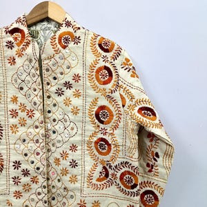 May include: An off-white jacket featuring intricate embroidery in shades of orange, red, and brown. The garment is adorned with circular floral patterns, vine-like motifs, and small reflective mirror accents throughout the front and sleeve panels.