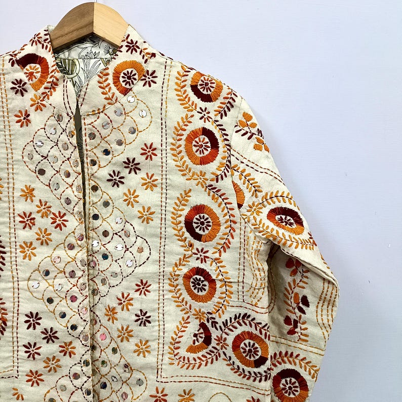 May include: An off-white jacket featuring intricate embroidery in shades of orange, red, and brown. The garment is adorned with circular floral patterns, vine-like motifs, and small reflective mirror accents throughout the front and sleeve panels.