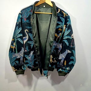 May include: Black bomber jacket with a vibrant jungle scene featuring zebras, flamingos, and lush foliage in teal, green, and yellow. The jacket has a green satin lining and ribbed cuffs and collar.