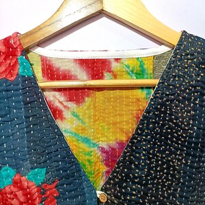 Op de afbeelding: Een patchwork vest met een V-hals ontwerp, hangend aan een houten hanger. Het vest heeft een mix van patronen, waaronder een bloemenprint, tie-dye en gestippelde stof. Het kleurenpalet omvat tinten blauw, rood, geel en wit.