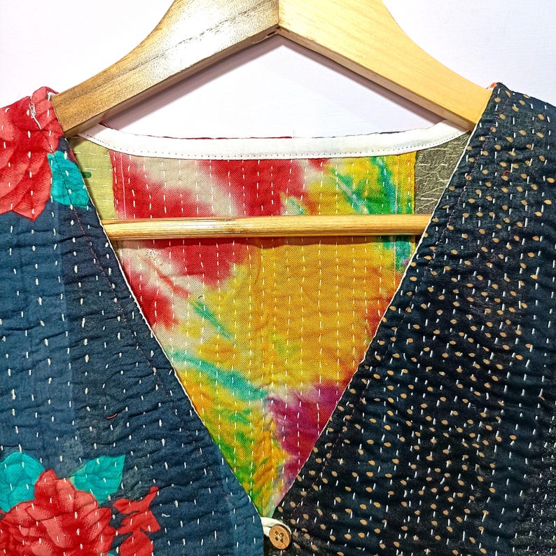 Op de afbeelding: Een patchwork vest met een V-hals ontwerp, hangend aan een houten hanger. Het vest heeft een mix van patronen, waaronder een bloemenprint, tie-dye en gestippelde stof. Het kleurenpalet omvat tinten blauw, rood, geel en wit.