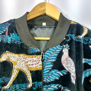 May include: Black velvet bomber jacket with a vibrant jungle print featuring a yellow and white leopard, blue foliage, and a parrot. The jacket has a dark green ribbed collar and a full-length zipper.
