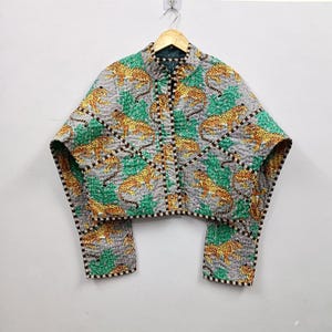 Pode incluir: Um casaco curto acolchoado com um padrão repetitivo de leopardos amarelos e pretos sobre um fundo cinzento com folhagem verde. A peça tem uma gola mandarim, mangas largas e bordas com riscas castanho-escuras e brancas.