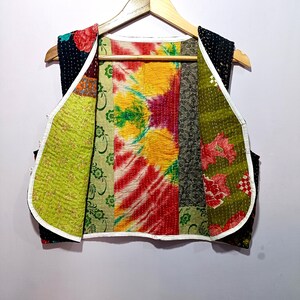 Op de afbeelding: Een kleurrijk patchworkvest met een verscheidenheid aan bloemen- en patroonstoffen. Het vest heeft een mix van groene, rode, gele en zwarte stoffen, met een witte bies. Het vest wordt gepresenteerd op een houten hanger.