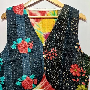 Op de afbeelding: Een omkeerbaar vest met een bloemenpatroon. De ene kant heeft een zwarte achtergrond met rode rozen en blauwgroene bladeren, de andere kant een zwart-blauw gestreept patroon met rode rozen. Het vest heeft een tie-dye voering en houten knopen.