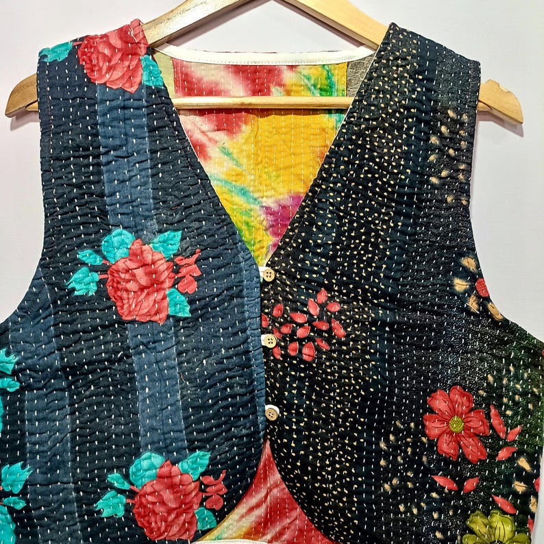 Op de afbeelding: Een omkeerbaar vest met een bloemenpatroon. De ene kant heeft een zwarte achtergrond met rode rozen en blauwgroene bladeren, de andere kant een zwart-blauw gestreept patroon met rode rozen. Het vest heeft een tie-dye voering en houten knopen.