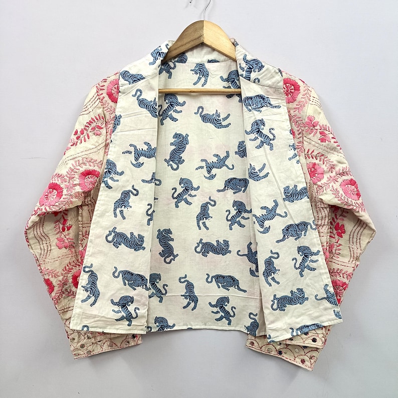 May include: A long-sleeve jacket featuring a cream-colored interior lining with a repeating blue tiger print. The exterior panels display a pink floral pattern on a cream background. The garment has an open front and a folded collar design.
