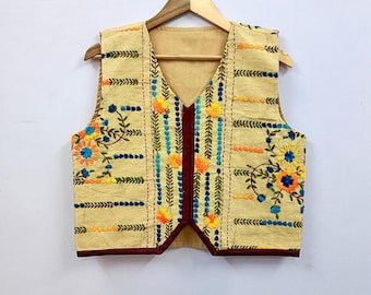 May include: A beige vest with colorful floral embroidery. The vest features a V-neck and is adorned with orange, blue, and yellow flowers, along with green leaves. The edges are trimmed with a dark red fabric. The vest is hanging on a wooden hanger.