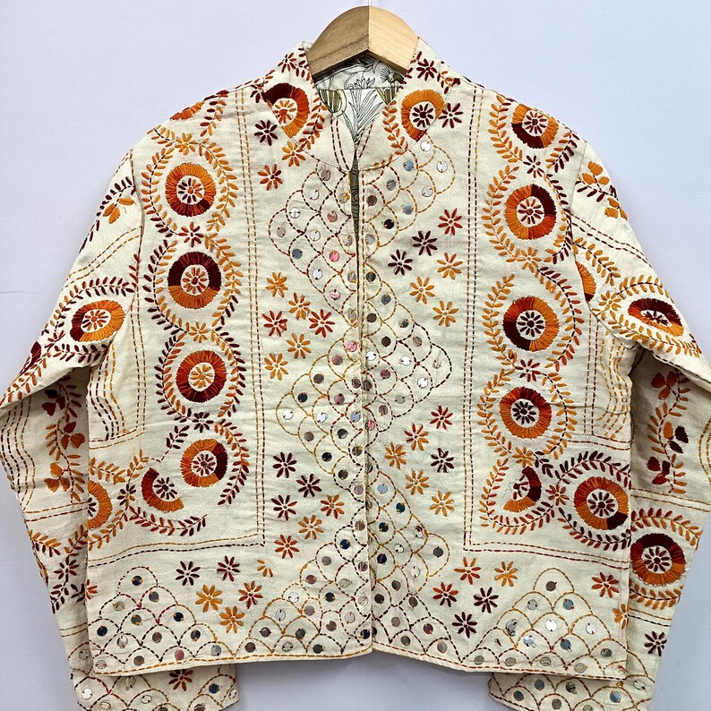 May include: An off-white long-sleeve jacket featuring intricate embroidery in shades of orange, red, and brown. The design includes circular floral patterns, small star-like motifs, and reflective mirror accents arranged in geometric grid layouts.