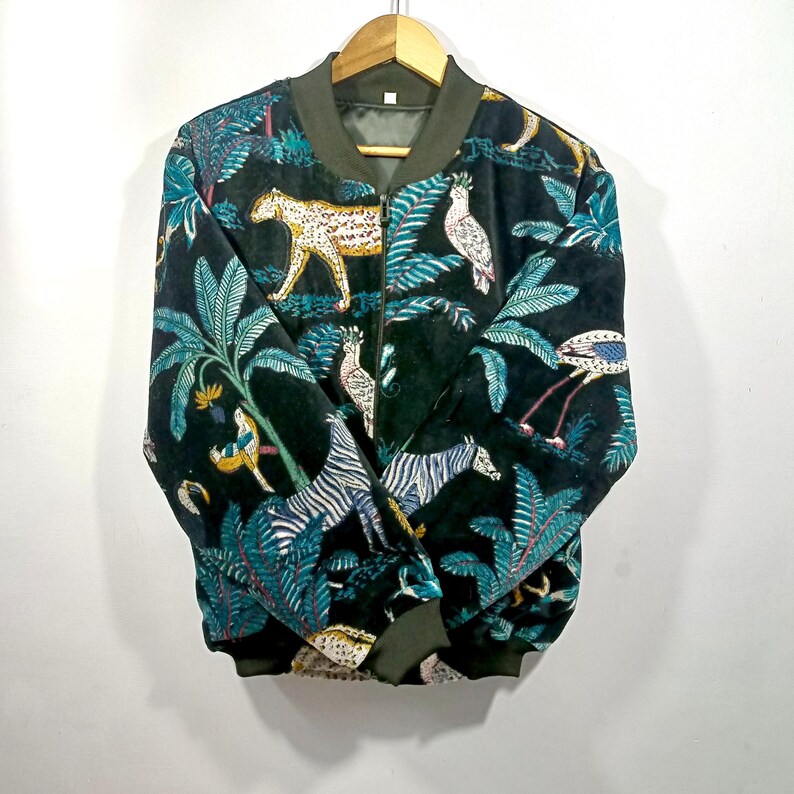 May include: Black bomber jacket with a jungle-themed pattern. The jacket features a repeating design of zebras, leopards, birds, and tropical plants in shades of teal, yellow, and white. The jacket has a green collar, cuffs, and hem.