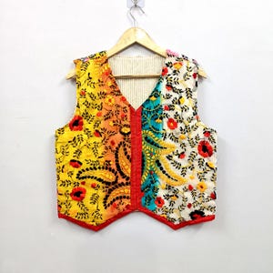 May include: A sleeveless waistcoat featuring a vibrant colour-blocked design with yellow, orange, and teal sections. The garment is adorned with intricate floral and paisley embroidery in red, yellow, and black thread, finished with a bright red trim.