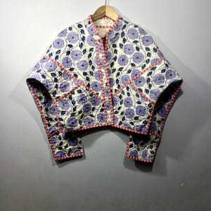 May include: A quilted jacket with a floral pattern in shades of purple and blue on an off-white background. The jacket has a mandarin collar, short sleeves, and is trimmed with red stitching. The jacket is hanging on a wooden hanger.