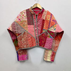 May include: A patchwork jacket in shades of pink, red, and orange, with a stand-up collar and long sleeves. The jacket is made from various fabric scraps, stitched together in a geometric pattern. The edges are trimmed with black and white striped fabric.