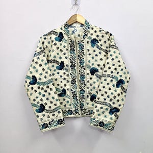 Könnte beinhalten: Cremefarbene Jacke mit floralem Muster. Die Jacke hat einen Stehkragen, lange Ärmel und einen Reißverschluss. Das florale Design umfasst blau und schwarz gestickte Blumen und Blätter, die einen dekorativen und detaillierten Look erzeugen. Die Jacke hängt an einem Holzbügel.