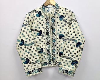 May include: Cream-colored jacket with a floral pattern. The jacket features a stand-up collar, long sleeves, and a zipper closure. The floral design includes blue and black embroidered flowers and leaves, creating a decorative and detailed look. The jacket is hanging on a wooden hanger.