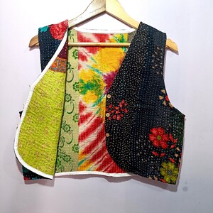 Op de afbeelding: Een patchwork vest met een verscheidenheid aan kleurrijke patronen en texturen. Het vest heeft een zwarte bloemenprint, een tie-dye ontwerp en groene bloempatronen. Het heeft een V-hals en is vastgezet met houten knopen. Het vest hangt aan een houten hanger.
