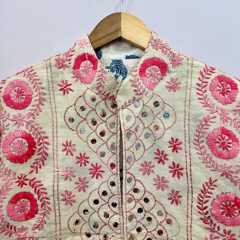 May include: An off-white jacket featuring vibrant pink circular floral embroidery and intricate scalloped patterns accented with small reflective mirrors. The garment has a mandarin collar and is displayed on a wooden hanger against a plain background.