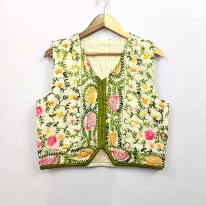 May include: Cream-coloured embroidered vest with a V-neck. The vest features a floral pattern with pink, yellow, and green embroidery. The edges are trimmed with green fabric. The vest is hanging on a wooden hanger.