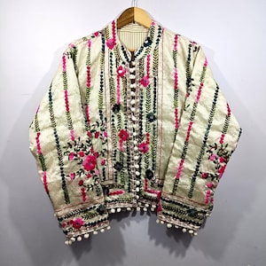 Peut inclure: Veste brodée crème avec des rayures verticales et des motifs floraux. La veste présente des broderies florales roses, vertes et noires, avec des pompons blancs le long des poignets et de l'ourlet. Elle a un devant boutonné et une doublure rayée.
