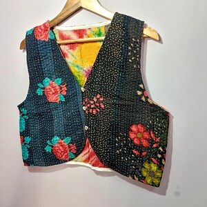 Op de afbeelding: Een patchwork vest met een V-hals ontwerp. Het vest heeft een mix van bloemen- en patroonstoffen in tinten zwart, blauwgroen, rood en geel. Het heeft een knoopsluiting en wordt weergegeven op een houten hanger.