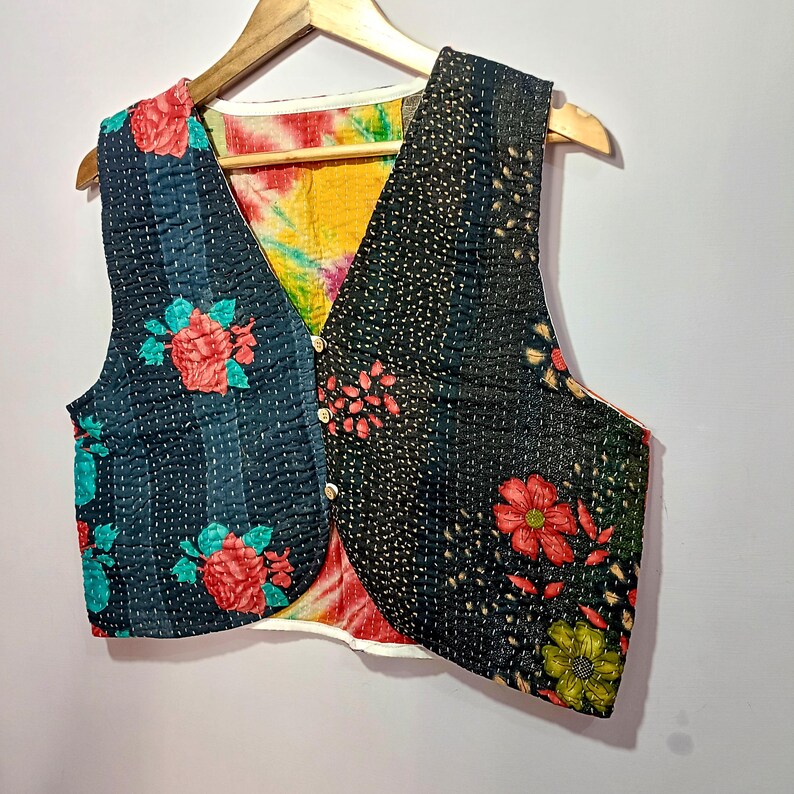 Op de afbeelding: Een patchwork vest met een V-hals ontwerp. Het vest heeft een mix van bloemen- en patroonstoffen in tinten zwart, blauwgroen, rood en geel. Het heeft een knoopsluiting en wordt weergegeven op een houten hanger.