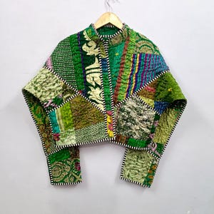 Pode incluir: Um casaco curto construído a partir de vários painéis de tecido patchwork verde com padrões e texturas intrincados. A peça inclui uma gola alta, mangas compridas e uma abertura frontal, tudo com acabamento em debrum às riscas pretas e brancas.